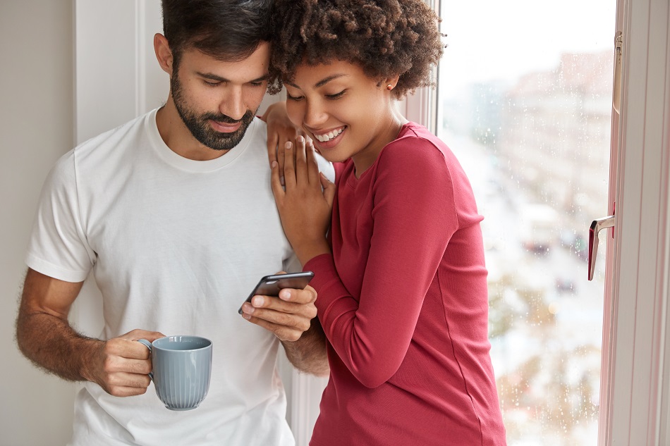 Image of mixed race friendly girl and boy wait for feedback from followers, develop their blog, stand closely near window, drink coffee, chat online, being addicted to smart phones, pose at home