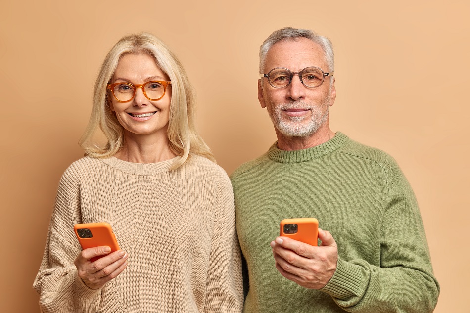 Horizontal shot of elderly couple use modern technologies hold smart phones reads text messages connected to wireless internet wear casual jumpers isolated over brown background. Retired pensioners