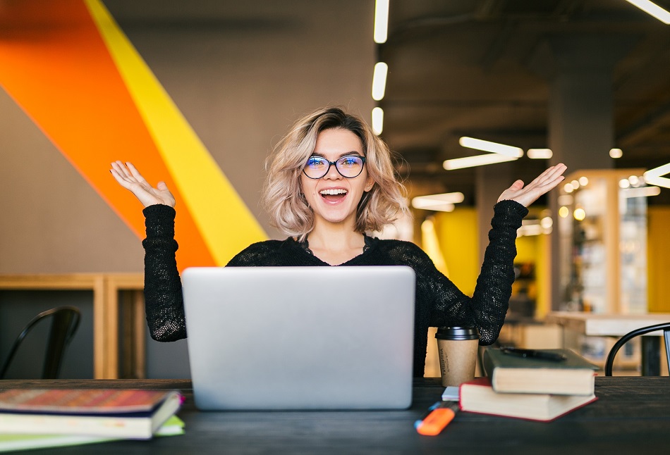 funny happy excited young pretty woman sitting at table in black shirt working on laptop in co-working office, wearing glasses